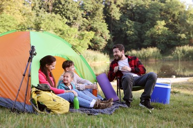 Photo of Happy family having break with camping tent, tourist equipment and drinks near river outdoors