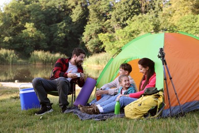 Photo of Happy family having break with camping tent, tourist equipment and drinks near river outdoors