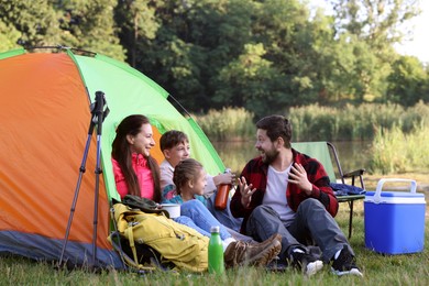 Photo of Happy family having break with camping tent, tourist equipment and drinks near river outdoors