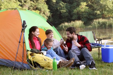 Photo of Happy family having break with camping tent, tourist equipment and drinks near river outdoors