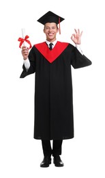 Photo of Young man in graduation gown and mortarboard with diploma showing ok gesture on white background