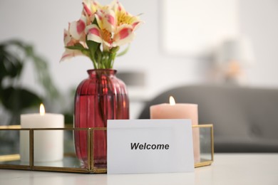 Photo of Card with word Welcome, vase with flowers and burning candles on white table indoors