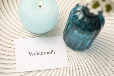 Photo of Card with word Welcome, burning candle and vase with flowers on white table, closeup