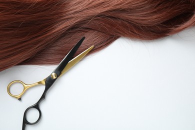 Photo of Auburn hair strand and hairdresser's scissors on white background, top view. Space for text