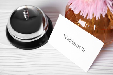 Photo of Card with word Welcome, desk bell and vase with flower on white wooden table, closeup