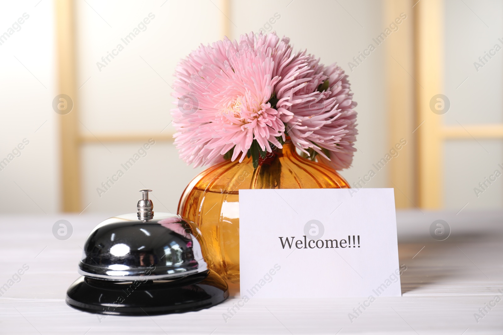 Card with word Welcome, desk bell and vase with flowers on white wooden table indoors Photo of Card with word Welcome, desk bell and vase with flowers on white wooden table indoors