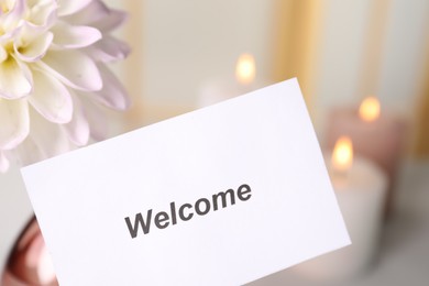 Photo of Card with word Welcome, flower and burning candles indoors, closeup