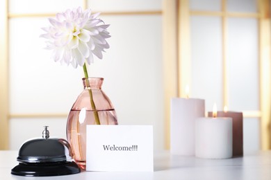 Photo of Card with word Welcome, desk bell, vase with flower and burning candles on white table. Space for text