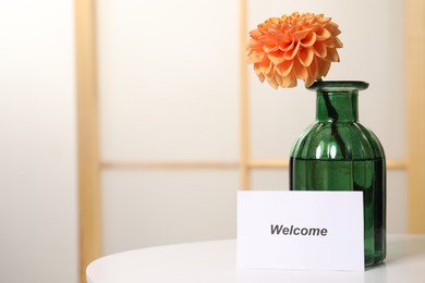 Photo of Card with word Welcome and vase with flower on white table indoors. Space for text