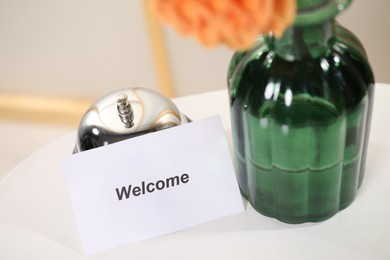 Photo of Card with word Welcome, desk bell and vase with flower on white table indoors, closeup