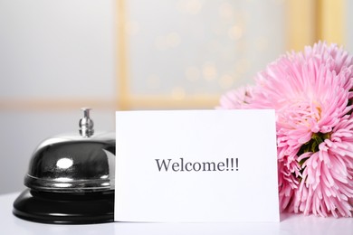 Photo of Card with word Welcome, desk bell and flowers on white table indoors, closeup