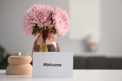 Photo of Card with word Welcome, vase with flowers and burning candle on white table indoors. Space for text