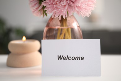 Photo of Card with word Welcome, vase with flowers and burning candle on white table indoors, closeup