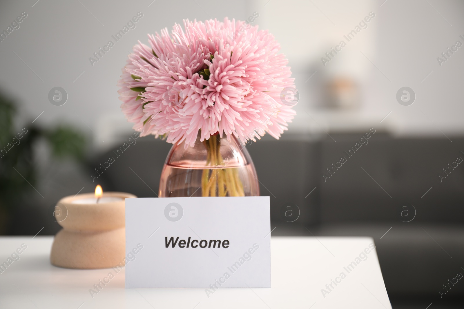 Card with word Welcome, vase with flowers and burning candle on white table indoors Photo of Card with word Welcome, vase with flowers and burning candle on white table indoors