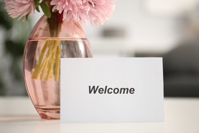 Photo of Card with word Welcome and vase with flowers on white table indoors, closeup