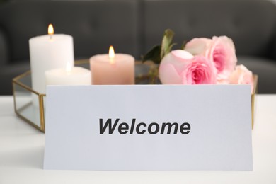 Photo of Card with word Welcome, roses and burning candles on white table, closeup