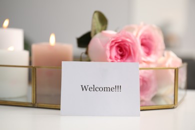 Photo of Card with word Welcome, roses and burning candles on white table, closeup