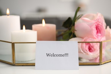 Photo of Card with word Welcome, roses and burning candles on white table, closeup