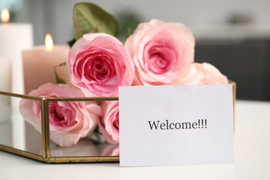 Photo of Card with word Welcome, roses and burning candles on white table, closeup