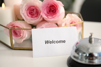 Photo of Card with word Welcome, desk bell and roses on white table, closeup