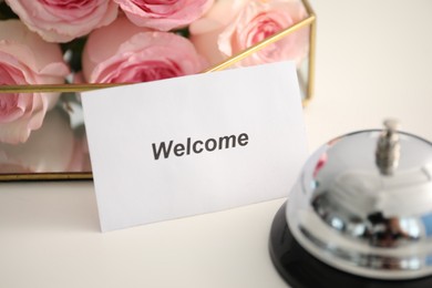 Photo of Card with word Welcome, desk bell and roses on white table, closeup