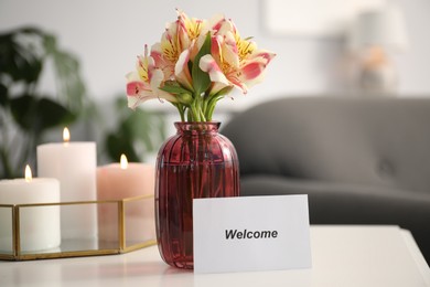 Photo of Card with word Welcome, vase with flowers and burning candles on white table indoors