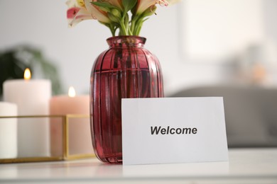 Photo of Card with word Welcome, vase with flowers and burning candles on white table indoors, closeup