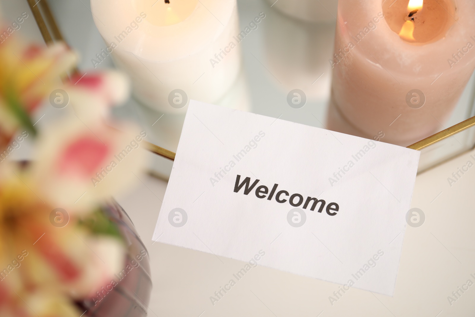 Photo of Card with word Welcome, vase with flowers and burning candles on white table, closeup