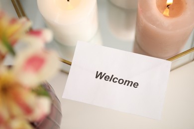 Photo of Card with word Welcome, vase with flowers and burning candles on white table, closeup