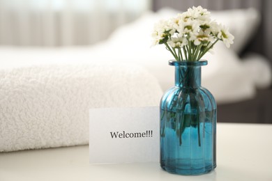 Photo of Card with word Welcome, vase with flowers and towel on white table indoors