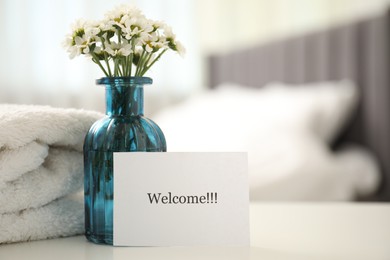 Photo of Card with word Welcome, vase with flowers and towel on white table indoors. Space for text