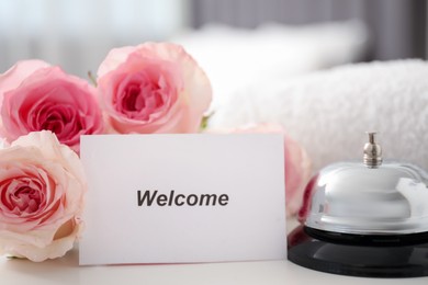 Photo of Card with word Welcome, desk bell, roses and towel on white table, closeup