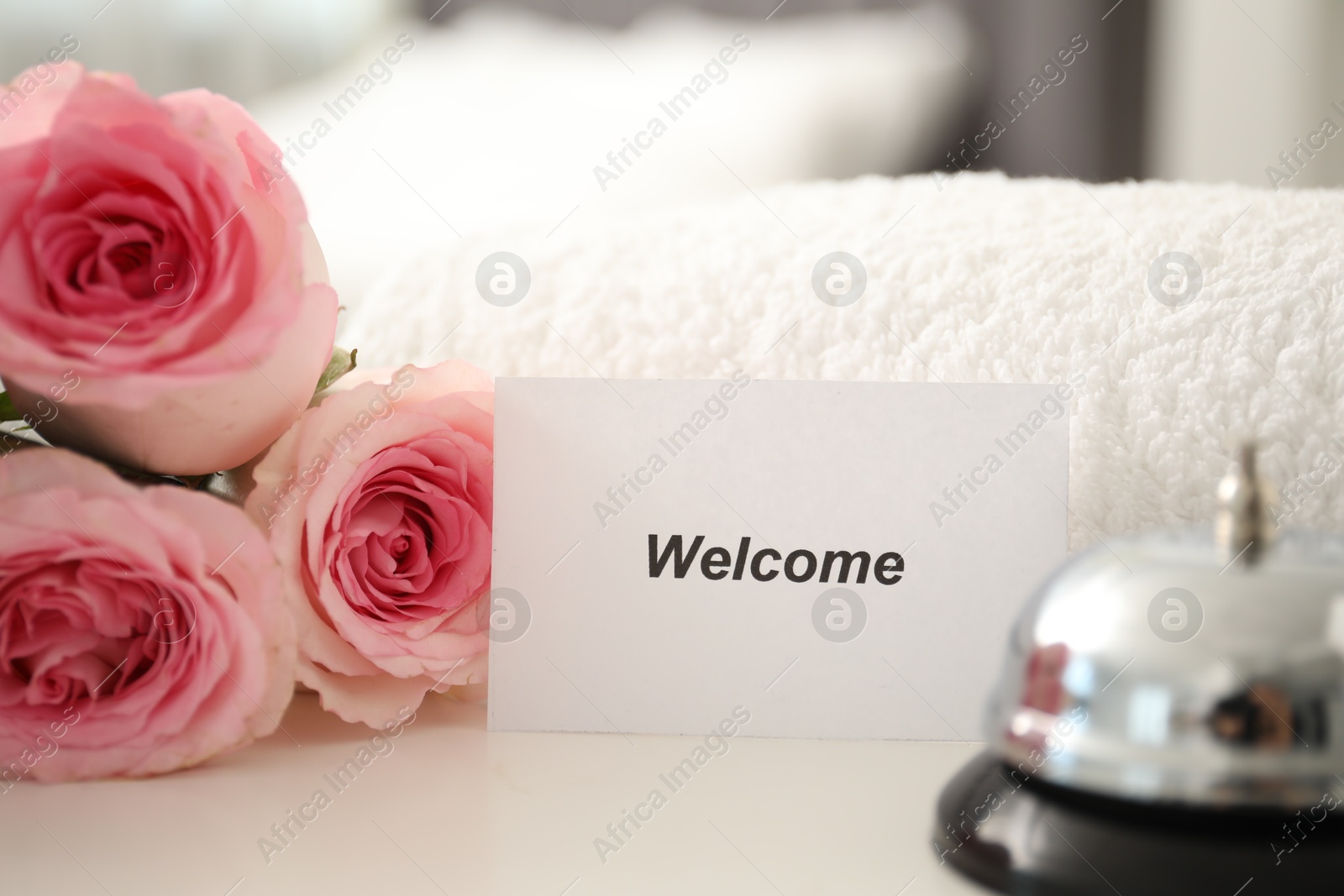 Photo of Card with word Welcome, desk bell, roses and towel on white table, closeup