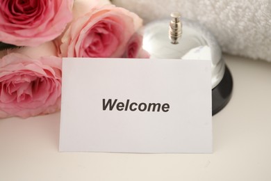 Photo of Card with word Welcome, desk bell, roses and towel on white table, closeup