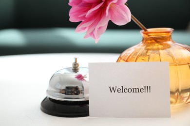 Photo of Card with word Welcome, desk bell and vase with flower on white table indoors, closeup