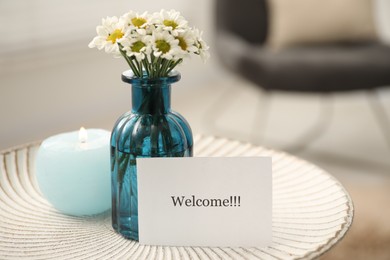 Photo of Card with word Welcome, burning candle and vase with flowers on white table indoors, closeup. Space for text