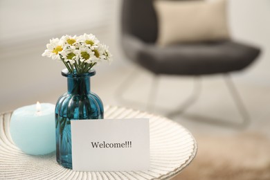 Photo of Card with word Welcome, burning candle and vase with flowers on white table indoors, closeup. Space for text