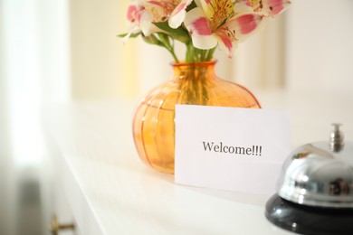 Photo of Card with word Welcome, desk bell and vase with flowers on white table indoors, closeup