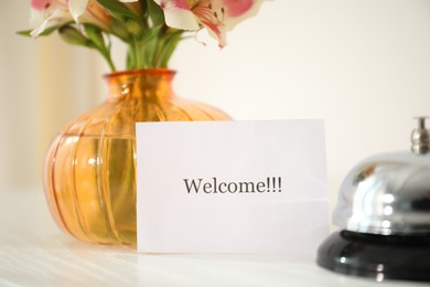 Photo of Card with word Welcome, desk bell and vase with flowers on white table indoors, closeup