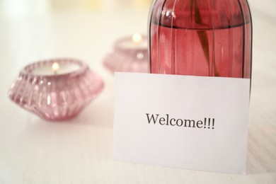 Photo of Card with word Welcome, vase with flower and candles on white table indoors, closeup