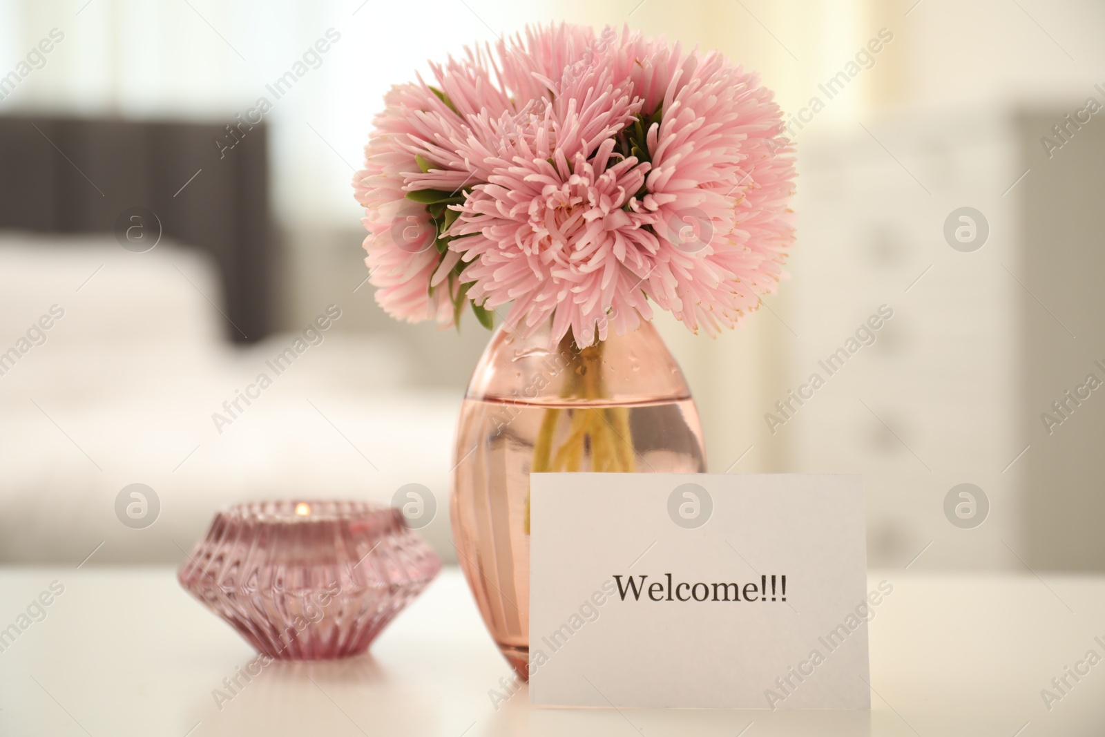 Photo of Card with word Welcome, vase with flowers and candle on white table indoors