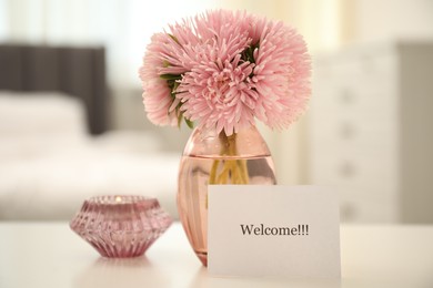 Photo of Card with word Welcome, vase with flowers and candle on white table indoors