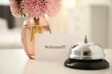 Photo of Card with word Welcome, desk bell and vase with flowers on white table indoors, closeup