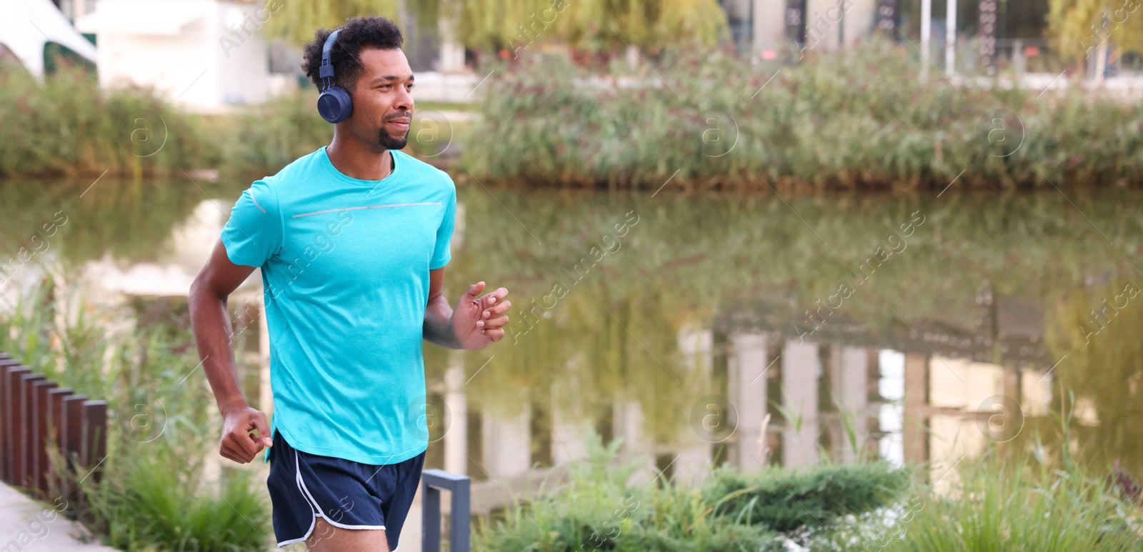 Photo of Handsome man in headphones running near river outdoors. Space for text