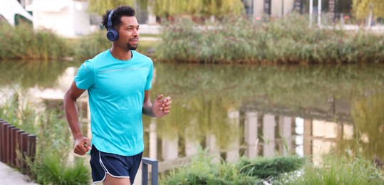 Photo of Handsome man in headphones running near river outdoors. Space for text
