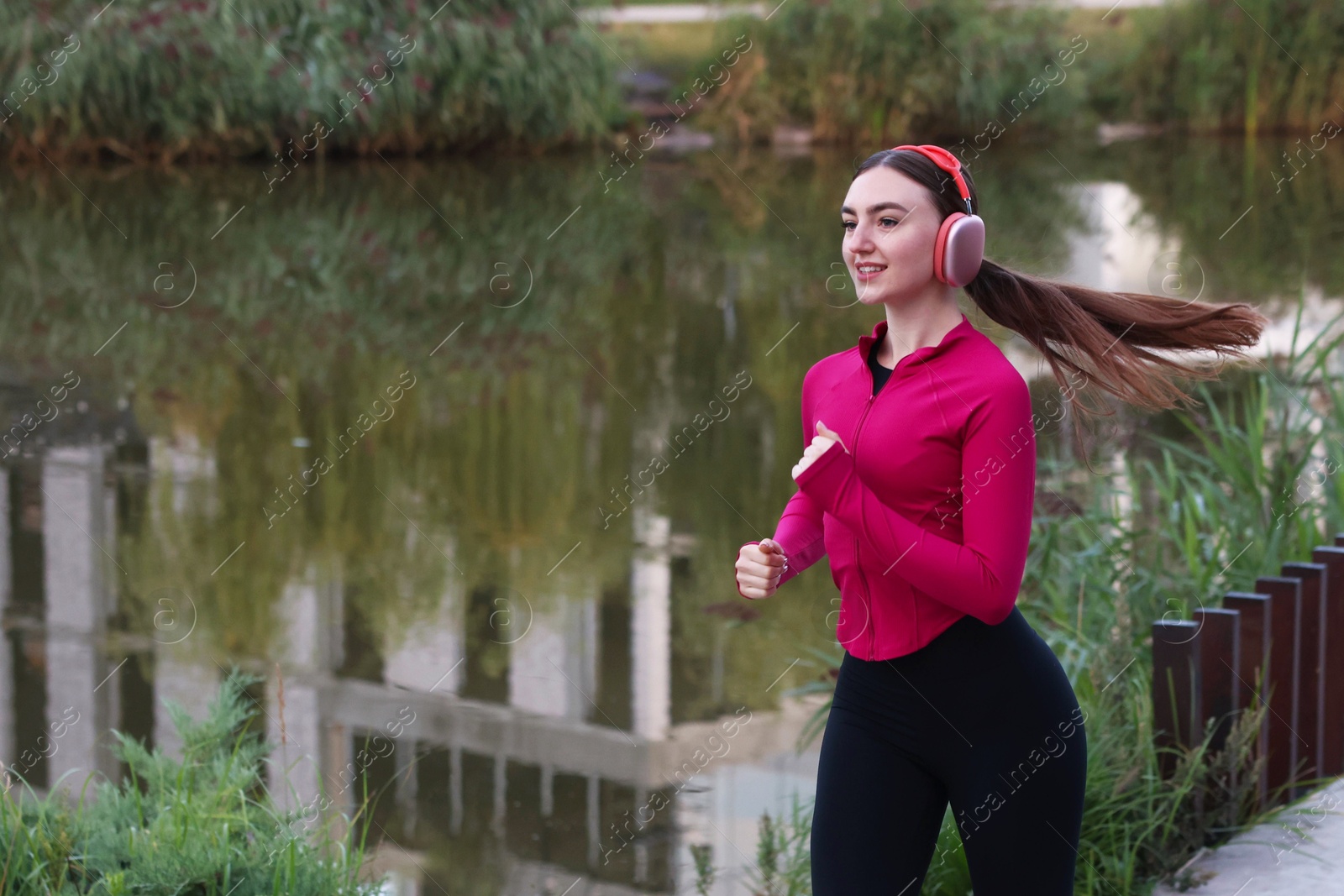 Photo of Smiling woman in headphones running near river outdoors. Space for text