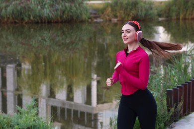 Photo of Smiling woman in headphones running near river outdoors. Space for text
