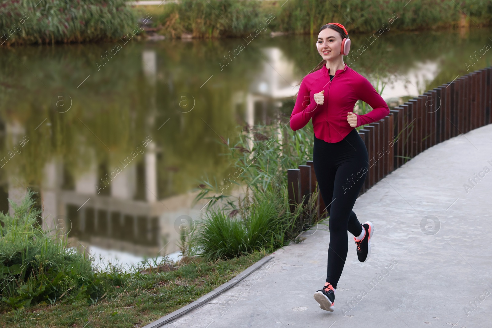 Photo of Smiling woman in headphones running near river outdoors. Space for text