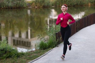 Photo of Smiling woman in headphones running near river outdoors. Space for text