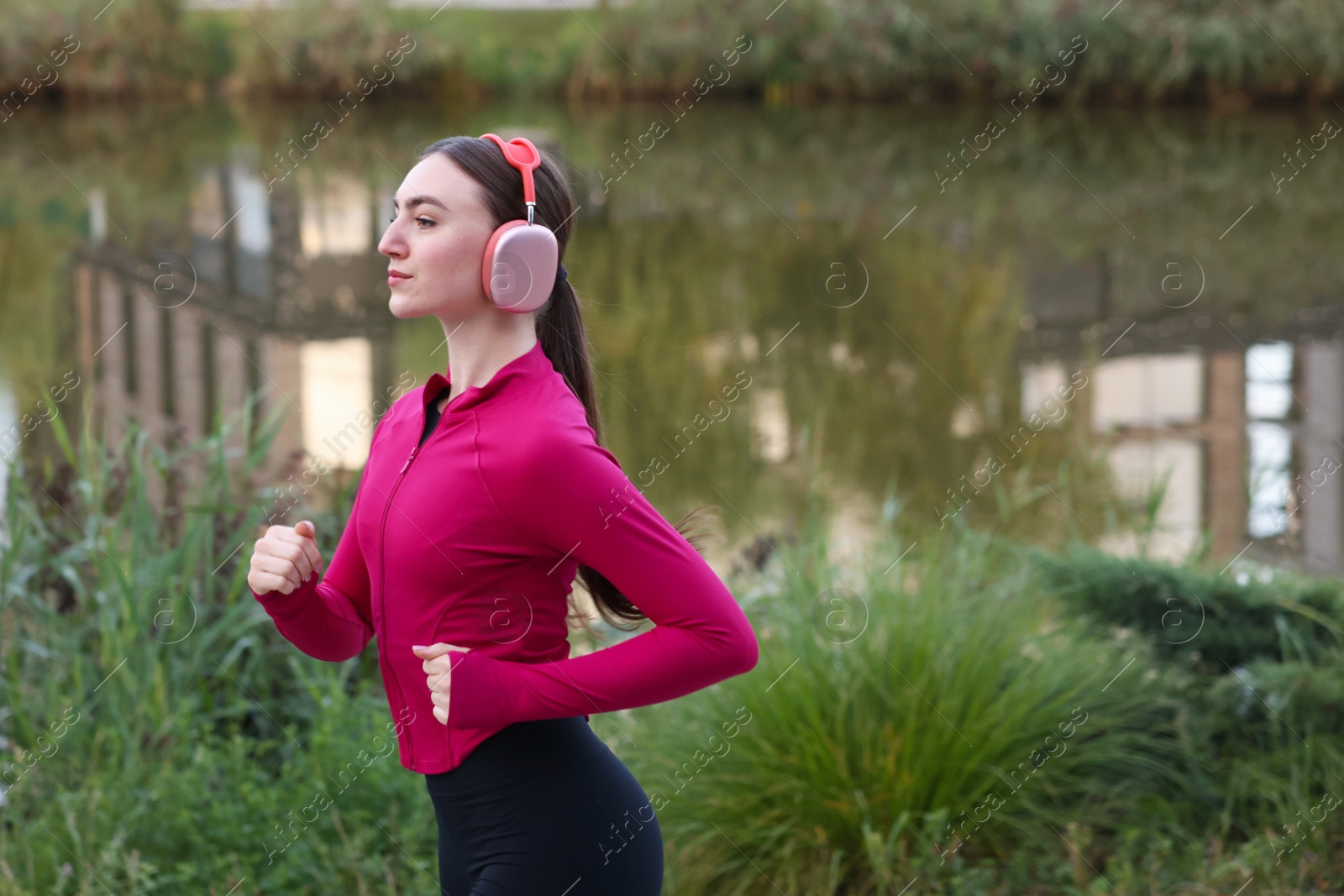 Photo of Beautiful woman in headphones running near river outdoors. Space for text